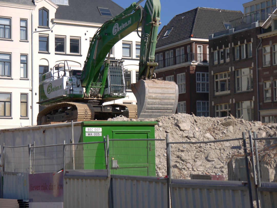Photo of a shovel on a concrete heap behind a metal construction fence. A large building is demolished to make space for new buildings. This demolition site is located close to Dam square. Street photography of construction works in old city Amsterdam by Fons Heijnsbroek; free download urban photo, The Netherlands. This street image I share in suitable high resolution for making a nice art print or wallpaper. / Gratis download foto van straatfotografie Amsterdam, Nederland: sloop werkzaamheden en bouwterrein aan het Rokin.